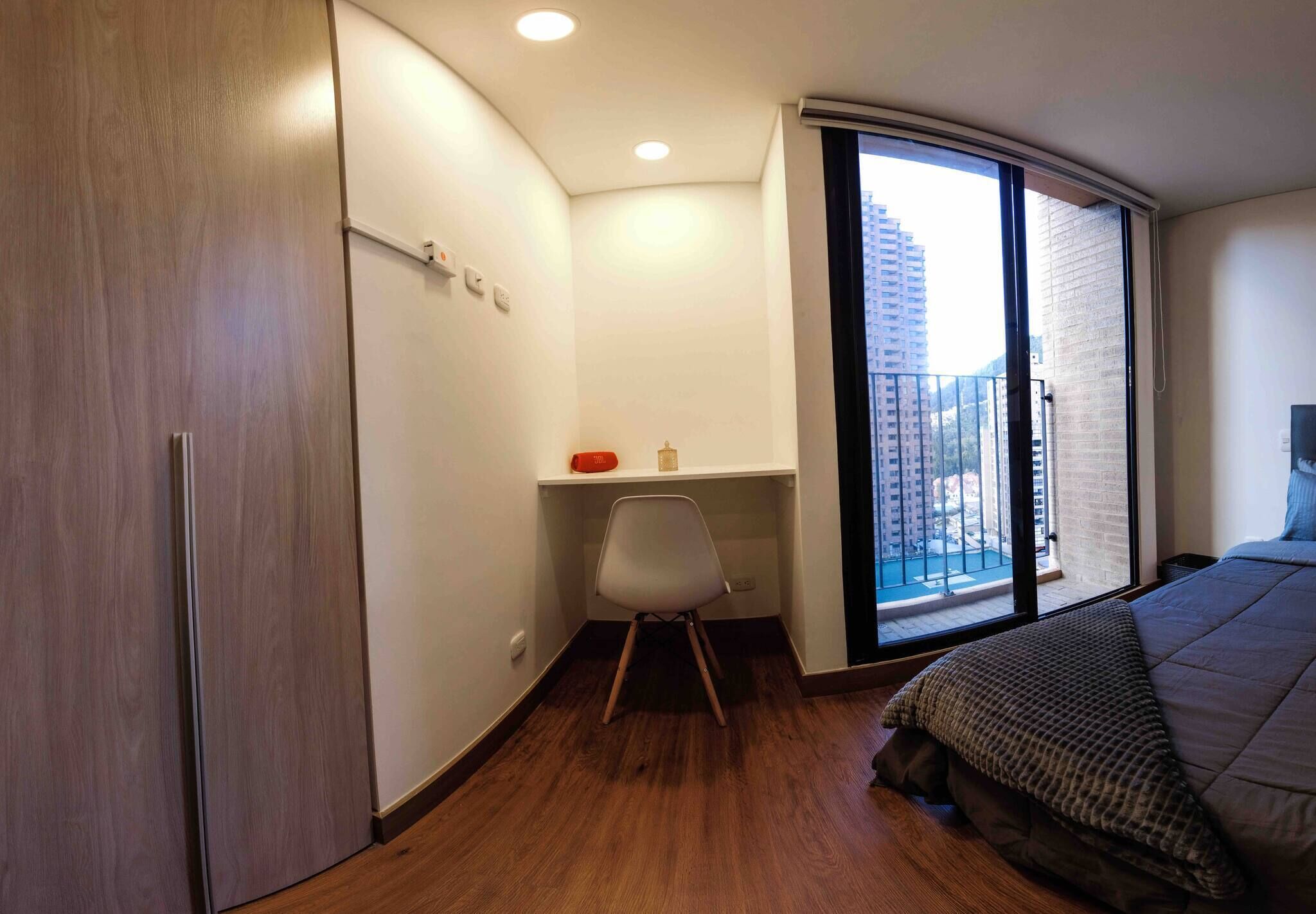 Un dormitorio con una cama, un escritorio con silla, un armario y un ventanal con vistas a edificios.