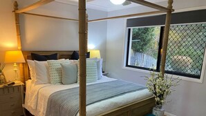 A four-poster bed with a wooden headboard, a bedside table with a lamp, a window with a view of greenery, and a blue bench. - Room-relax & Unwind on the Gold Coast (Gold Coast)