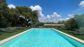 Una piscina rectangular con agua azul cristalina, rodeada de césped, sillas de descanso y árboles.