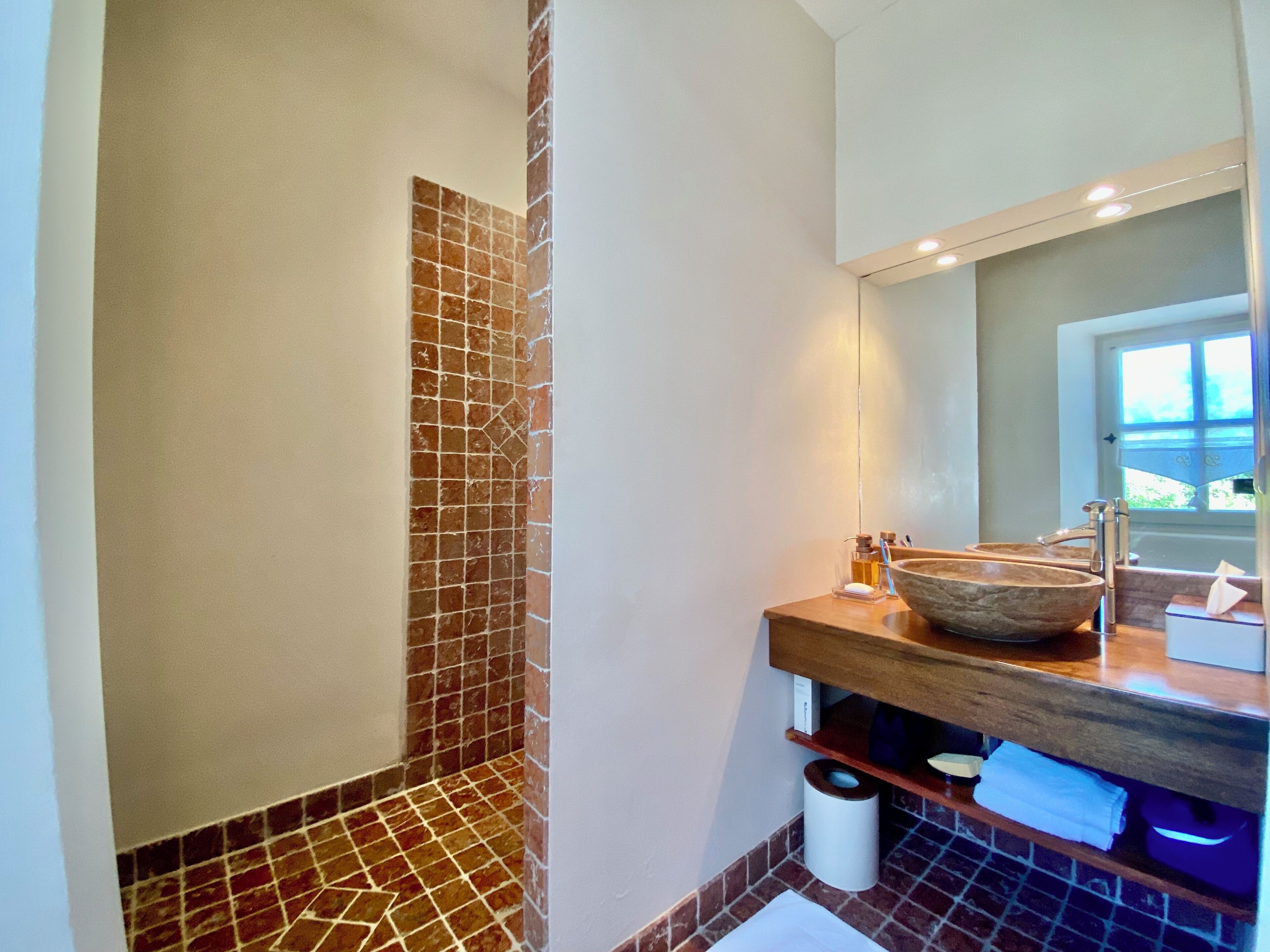 Une salle de bain dotée d’un grand miroir, d’un meuble-lavabo en bois, d’un évier en pierre et d’une zone de douche carrelée.