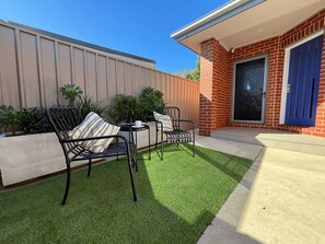 An outdoor patio with two metal chairs and a small table, surrounded by green artificial grass, against a brick wall and a building with a blue door.