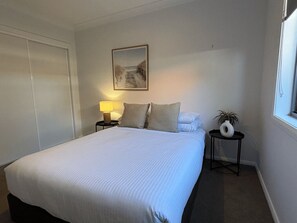 A neatly made bed with a white duvet, a bedside lamp, a small table with a vase, and a framed picture on the wall.