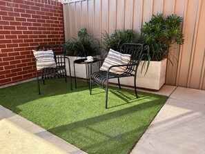 Une terrasse avec deux chaises en métal et une petite table, entourée de plantes en pot et d’un mur en briques.