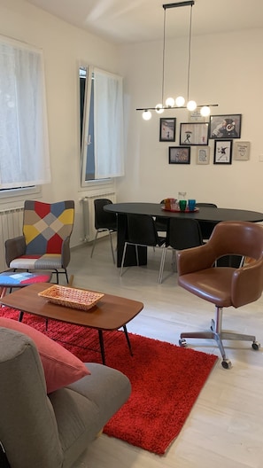 Una sala de estar con sofá, una mesa de centro, un tapete rojo, un sillón estampado, una mesa de comedor con sillas y decoración en la pared.