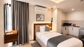 A hotel room with a bed, bedside table, curtains, an air conditioning unit, a framed picture, a kitchen area with a microwave, and a refrigerator.