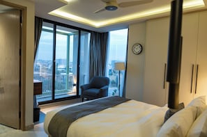 Une chambre d’hôtel avec un lit, une chaise, une fenêtre donnant sur des immeubles, une horloge au mur et un ventilateur de plafond.