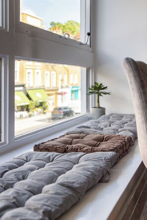 Una cama con un edredón gris, una ventana con vistas a edificios, y una planta en maceta en el alféizar.