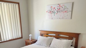 Une chambre à coucher avec un lit en bois, du linge de lit blanc, une serviette rose, une table de chevet et une commode, ainsi qu’un tableau représentant des flamants roses.