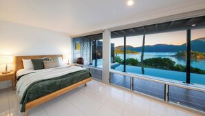 Ein modernes Schlafzimmer mit einem großen Bett, einem Nachttisch, einer Lampe und Blick auf einen See und Berge durch große Fenster.