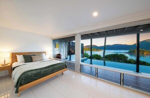 Ein modernes Schlafzimmer mit einem großen Bett, einem Nachttisch, einer Lampe und Blick auf einen See und Berge durch große Fenster.