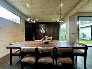A modern dining area with a large rectangular table, two pendant lights, and a view of a garden through a glass door. - Seseh House by Alfred in Bali (Cemagi)