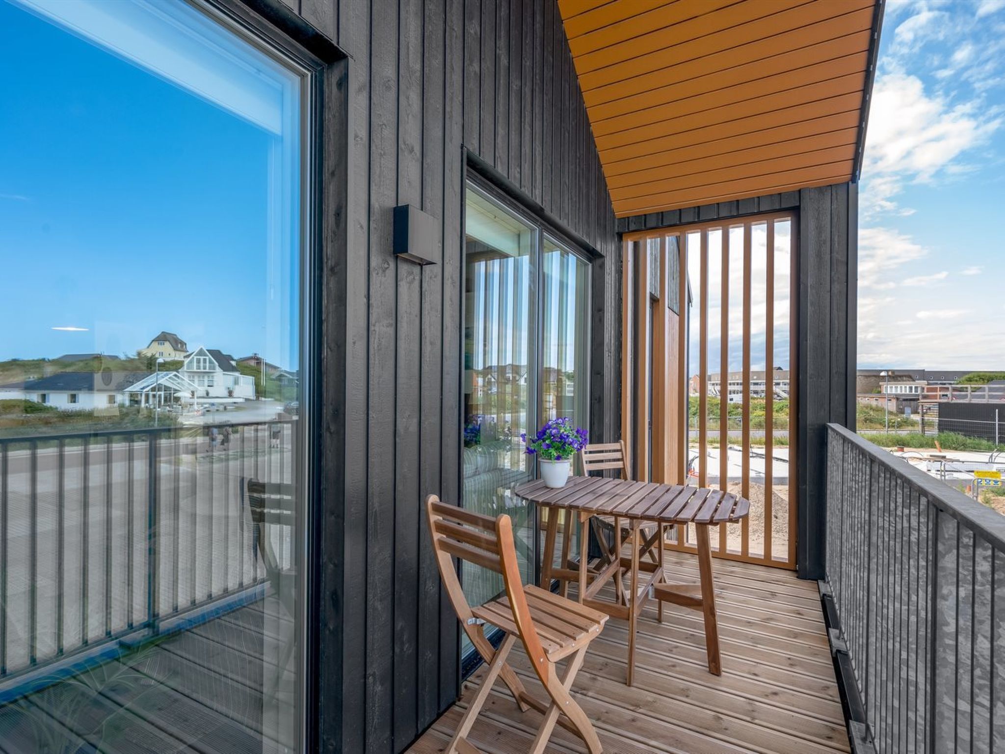 Uma varanda com mesa e cadeiras de madeira, um vaso de planta e vista para casas ao longe.