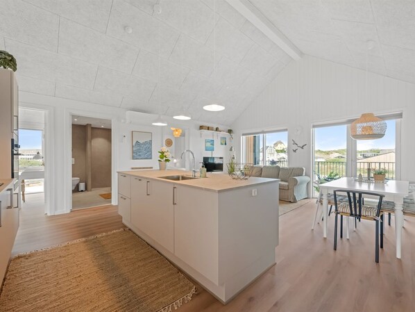 Une cuisine moderne dotée d’un îlot central, un coin repas avec une table et des chaises, et un espace de vie avec un canapé et une vue sur l’extérieur.