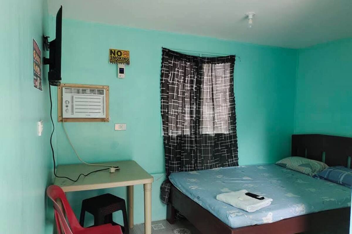 A room with teal walls, a bed with a blue mattress, a desk with a chair, a television, and a 'No Smoking' sign.