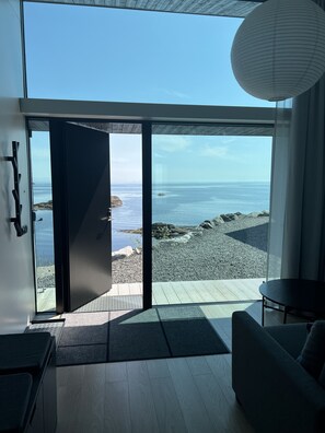 Et moderne interiør med en stor glasdør, der fører til udsigt over stranden, trægulv og en mørk sofa.
