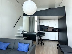 Una sala de estar moderna con un sofá gris y cojines azules, una cocina con armarios blancos y encimeras negras, y un gran farol de papel blanco colgado del techo.