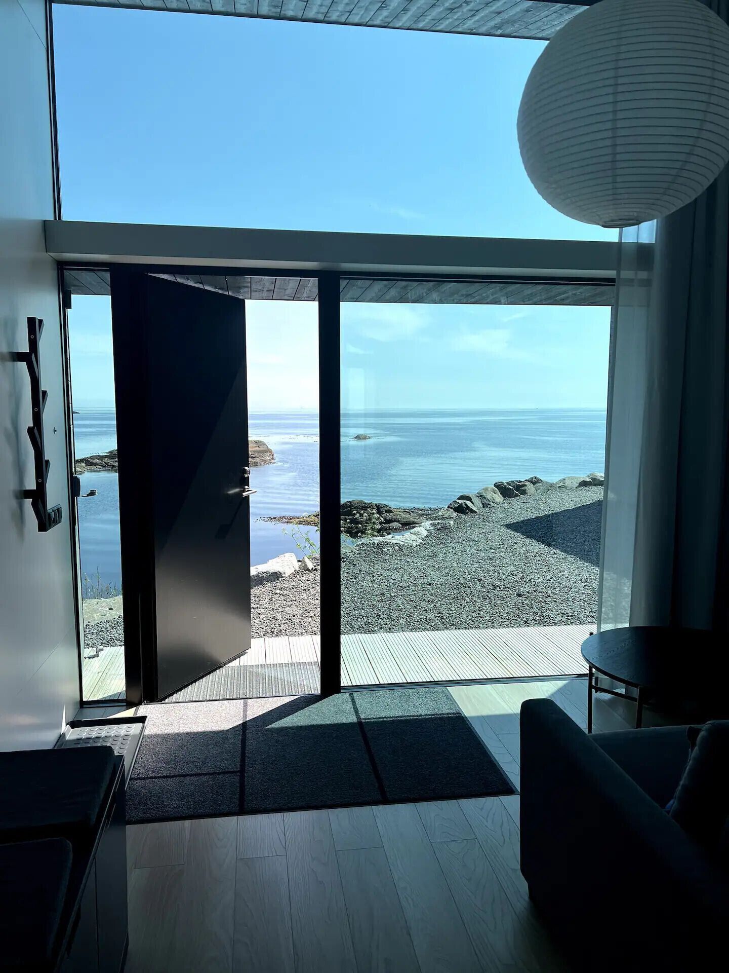 En modern inredning med en stor glasdörr som leder ut till en terrass med utsikt över stranden, ett trägolv och en enkel sittgrupp.