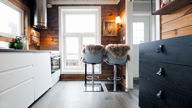 A kitchen with wooden walls, white cabinets, two bar stools with fur-cushioned seats, a black dresser, and a window with a view.