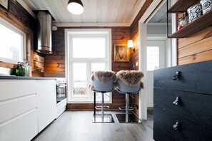 A kitchen with wooden walls, white cabinets, two bar stools with fur-cushioned seats, a black dresser, and a window with a view.