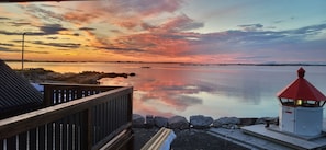 A waterfront view with a wooden deck, a small lighthouse, and calm water reflecting the sky.