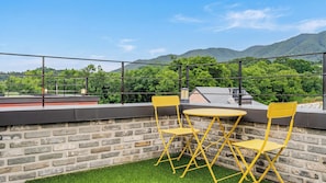 Una terraza en la azotea con un muro de ladrillo, dos sillas amarillas, una mesa redonda pequeña y una barandilla de metal.