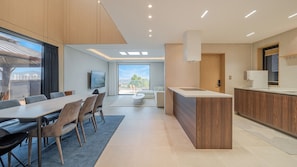 Una cocina moderna con una isla central, un comedor con mesa y sillas, y una sala de estar con sofá y vistas al exterior.