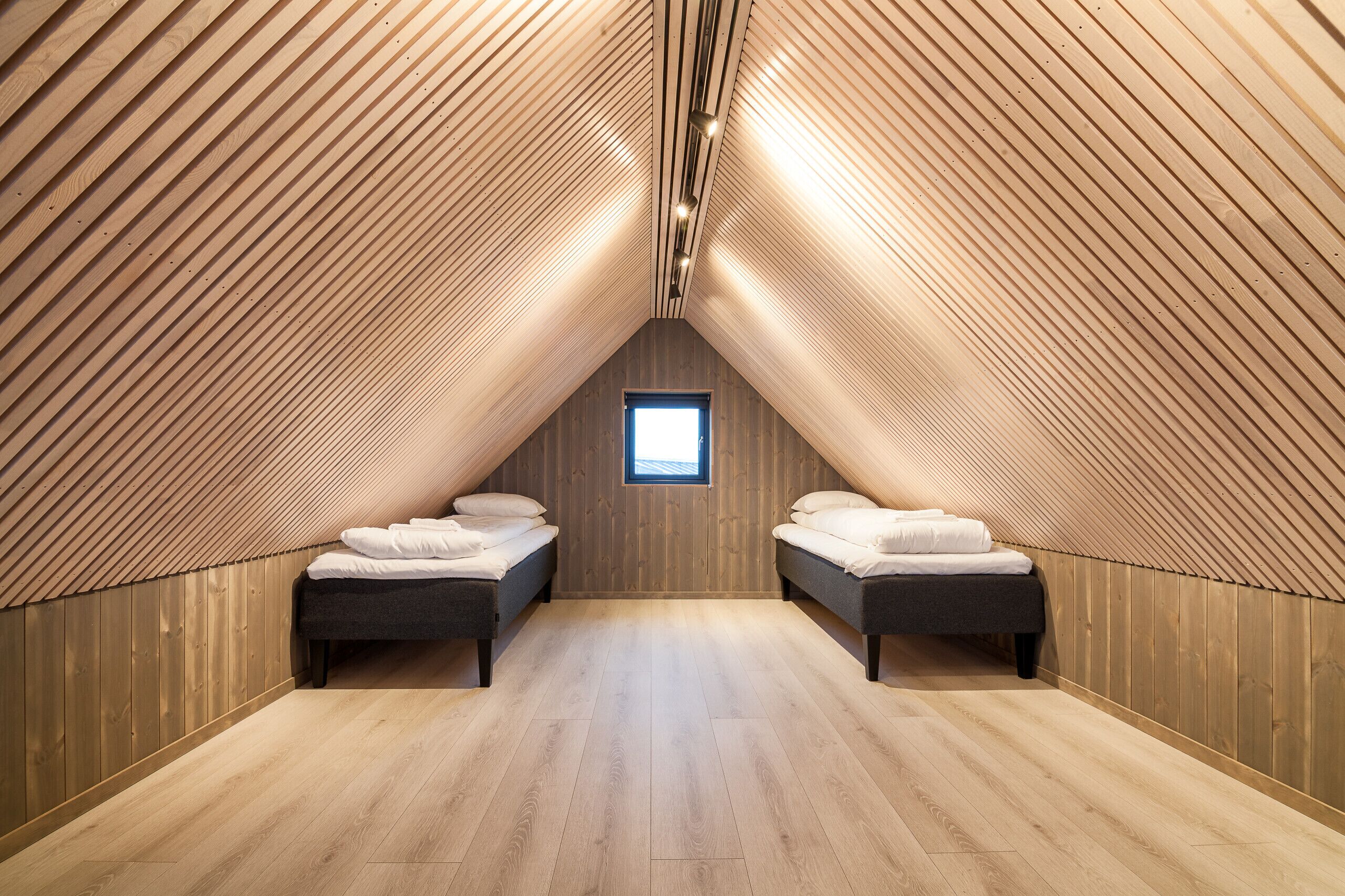 Une chambre mansardée avec des lambris en bois au plafond, deux lits, une petite fenêtre et un plancher en bois.