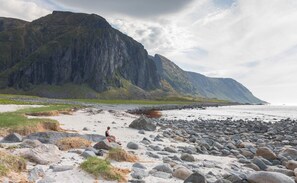 Une plage rocheuse où l’on voit une personne assise sur un rocher, avec des montagnes en arrière-plan et un ciel nuageux.
