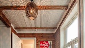 Una habitación acogedora con techo de madera, un sofá con cojines de colores, una mesa con lámpara y una puerta que da a otra habitación.