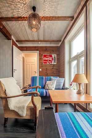 Une pièce chaleureuse avec un plafond en bois, un canapé agrémenté de coussins colorés, une table avec une lampe et une porte donnant sur une autre pièce.