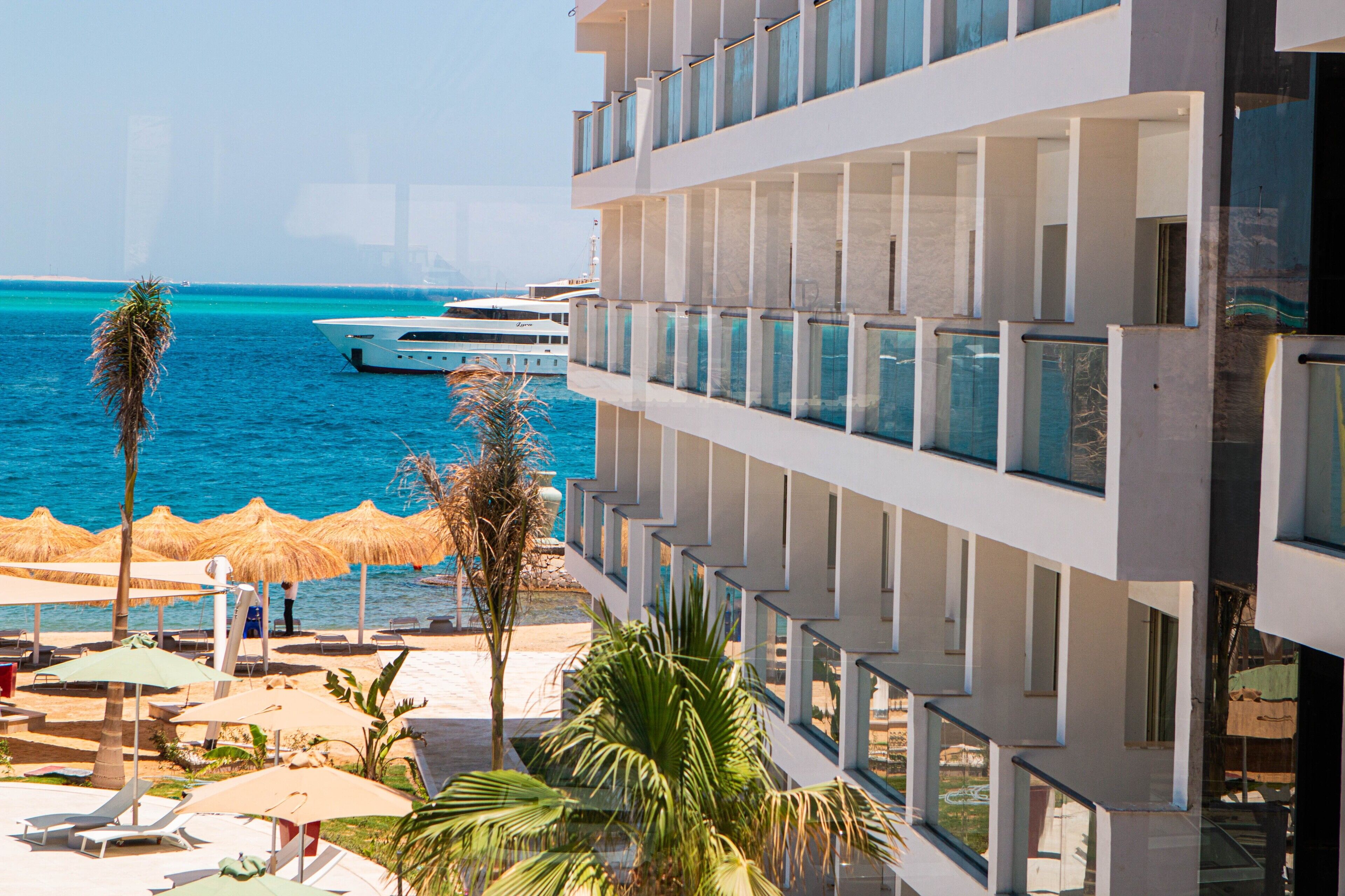 Un hotel con un balcone affacciato su una spiaggia, una zona piscina con lettini e uno yacht in acqua.