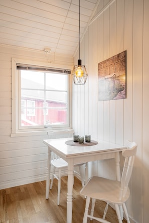 Une pièce avec une table blanche, deux chaises, un luminaire suspendu, une fenêtre donnant sur un immeuble rouge et un tableau accroché au mur.
