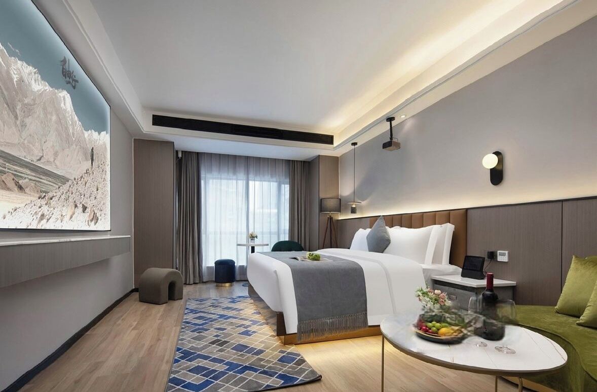 Ett modernt hotellrum med en säng, ett sängbord, en lampa och ett stort fönster.
