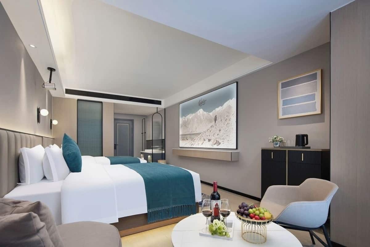 Una habitación de hotel moderna con una cama, un área de descanso, una mesa de comedor con fruta y vino, y un cuadro grande en la pared.