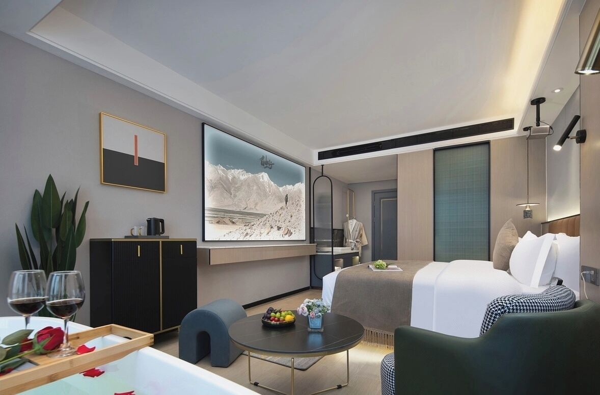 A modern hotel room with a bed, a dining area, a TV, and a large window.
