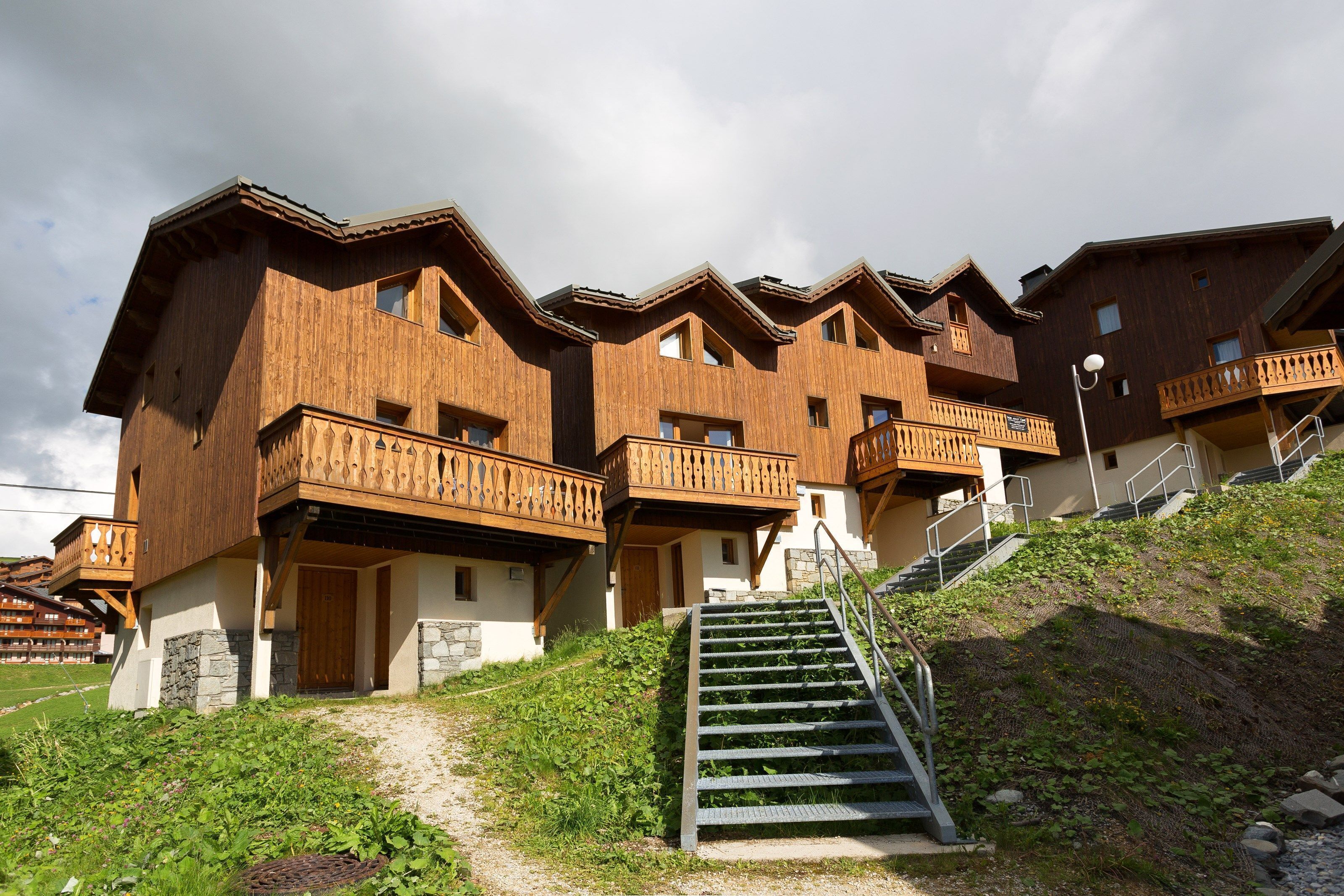 A row of wooden chalet-style buildings with balconies, a staircase leading up to the entrance, and a grassy slope in the foreground.