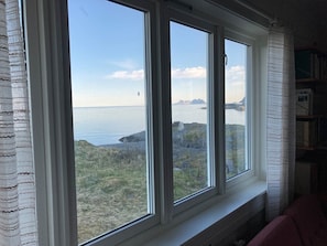 A room with a large window overlooking a coastal landscape, a bookshelf, and a red chair. - House with amazing view & location in Tind (Moskenes)