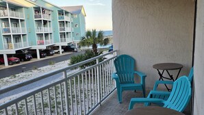 A balcony with turquoise chairs and a table, overlooking a beach and a row of light blue buildings.