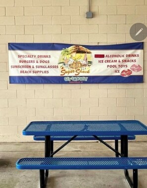 Bannerissa mainostetaan rantakahvilaa, jossa on tarjolla erikoisjuomia, hampurilaisia, hot dogeja, aurinkorasvaa, aurinkolaseja, jäätelöä, uima-allasleluja ja jäätä.