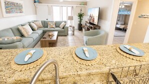 A living room with a sofa, a coffee table, a TV, and a kitchen area with granite countertops and a sink.