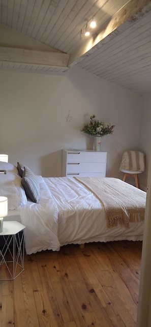 Een slaapkamer met een houten vloer, een bed met witte beddengoed, een klein wit dressoir, een nachtkastje met een lamp en een stoel.