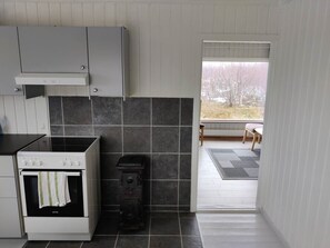 A kitchen with a stove, oven, and a small appliance, a window with a view of trees, and a door leading to an outdoor area.
