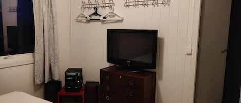 A bedroom with a bed, a TV on a wooden dresser, a coat rack, and a small table with electronic devices.