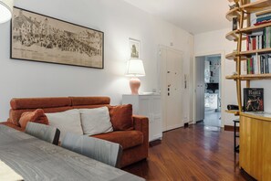 Ein Wohnzimmer mit einer Couch, einer Lampe, einem Bücherregal und einem gerahmten Bild an der Wand.