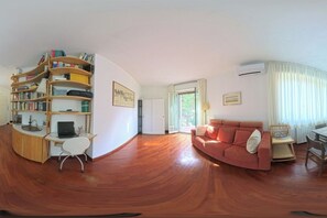 Ein Zimmer mit Holzboden, der leicht gewölbt ist, einem roten Sofa, einem weißen Schreibtisch mit Laptop, einem Bücherregal und einem großen Fenster mit Vorhängen.