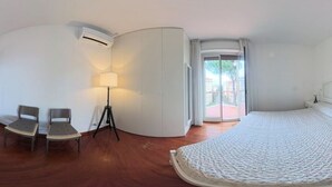 Une chambre d’h ôtel avec une armoire blanche aux lignes courbes, un lit recouvert d’une couvre-lit à motifs, deux chaises, une lampe sur pied et une vue sur l’extérieur par une porte vitrée.