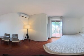 Ein Hotelzimmer mit einem gebogenen, weißen Kleiderschrank, einem Bett mit gemustertem Bettbezug, zwei Stühlen, einer Stehlampe und Blick durch eine Glastür nach draußen.