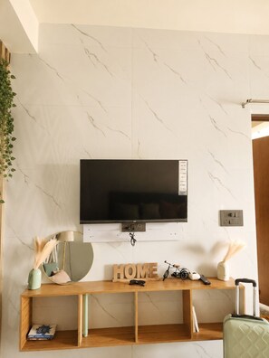 A modern living room with a TV mounted on a marble wall, a wooden console table with a 'HOME' sign, and a green suitcase.