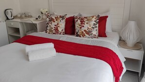 A neatly made bed with a red blanket, a white pillow, and a floral pillow. There is a white nightstand with a lamp and a small table with a kettle and cups.