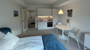Habitación de hotel compacta con cama, pequeña kitchenette, mesa de comedor con sillas y baño visible a través de una puerta abierta.